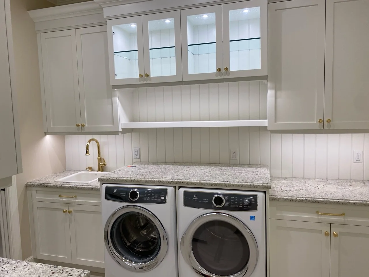 White laundry room cabinetry with illuminated glass display cabinets and gold hardware